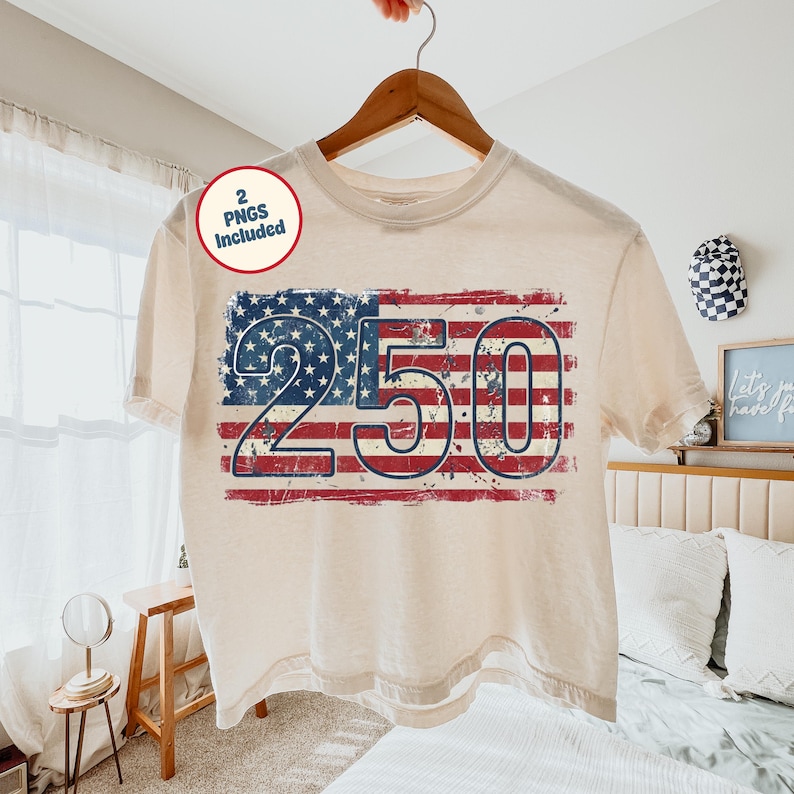 Puede incluir: Camiseta color crema con un dise&ntilde;o desgastado de la bandera estadounidense y el n&uacute;mero "250". La camiseta est&aacute; colgada de una percha de madera. Una peque&ntilde;a pegatina circular con el texto "2 PNGs Included" es visible.