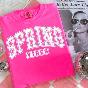 May include: A bright pink sweatshirt with the words "SPRING VIBES" in white block letters. The letters are filled with a floral pattern of pastel-colored flowers. The sweatshirt is folded on a pink and white checkered surface.