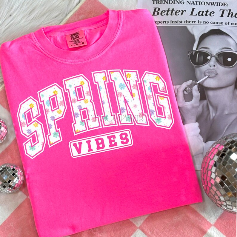 May include: A bright pink sweatshirt with the words "SPRING VIBES" in white block letters. The letters are filled with a floral pattern of pastel-colored flowers. The sweatshirt is folded on a pink and white checkered surface.