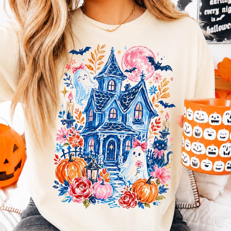 May include: A cream-colored crewneck sweatshirt featuring a colorful graphic print of a blue haunted house, two ghosts with pink bows, orange pumpkins, red roses, bats, a pink moon, and lit candles. The design is centered on the front of the garment.