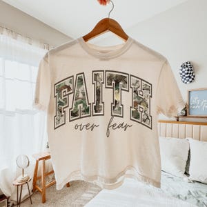 May include: A cream-colored t-shirt with the words "FAITH over fear" printed on the front. The word "FAITH" is in a block letter style with a camouflage pattern. The words are in a dark gray font.