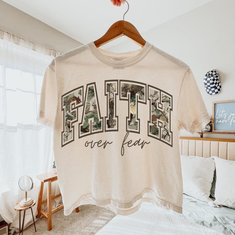 May include: A cream-colored t-shirt with the words "FAITH over fear" printed on the front. The word "FAITH" is in a block letter style with a camouflage pattern. The words are in a dark gray font.
