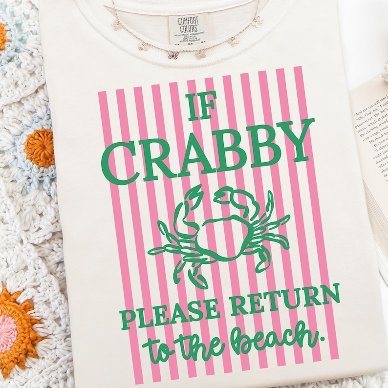 Puede incluir: Camiseta color crema con el texto "IF CRABBY PLEASE RETURN to the beach." El dise&ntilde;o presenta una ilustraci&oacute;n de cangrejo verde y rayas verticales rosas. Se ve un delicado collar con peque&ntilde;os dijes.