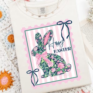 May include: Cream-colored sweatshirt featuring a floral patterned bunny graphic with the words "Happy Easter" in black script. The bunny is set within a pink and green frame with a pink bow. A delicate butterfly necklace is visible.