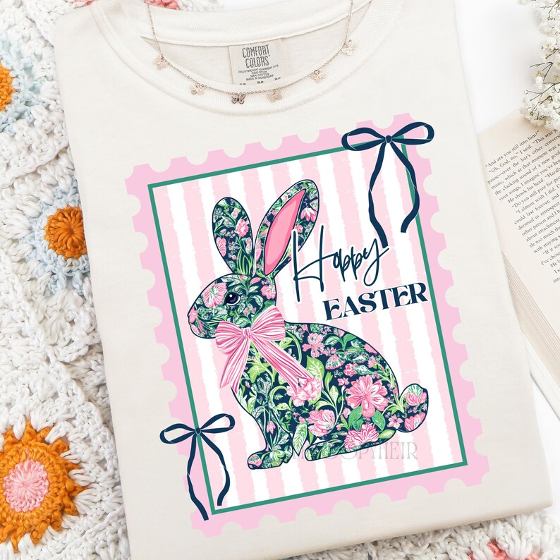 May include: Cream-colored sweatshirt featuring a floral patterned bunny graphic with the words "Happy Easter" in black script. The bunny is set within a pink and green frame with a pink bow. A delicate butterfly necklace is visible.