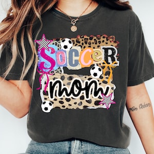 May include: Dark gray t-shirt with a colorful "Soccer Mom" graphic. The design features the words "Soccer Mom" in a collage style with soccer balls, stars, and leopard print accents. A casual, comfortable tee.