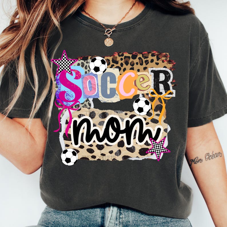 May include: Dark gray t-shirt with a colorful "Soccer Mom" graphic. The design features the words "Soccer Mom" in a collage style with soccer balls, stars, and leopard print accents. A casual, comfortable tee.