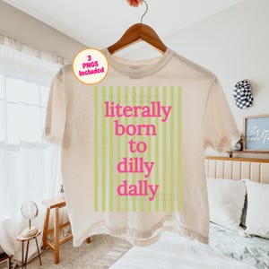 May include: A cream-colored t-shirt with the text "literally born to dilly dally" in pink letters, set against a background of vertical green stripes. The shirt is hanging on a wooden hanger.