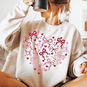 May include: Cream-colored sweatshirt featuring a heart design made of pink and red bows and hearts. The bows have intricate details, and the hearts are solid. The sweatshirt is a casual, comfortable style.