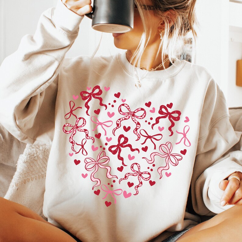May include: Cream-colored sweatshirt featuring a heart design made of pink and red bows and hearts. The bows have intricate details, and the hearts are solid. The sweatshirt is a casual, comfortable style.