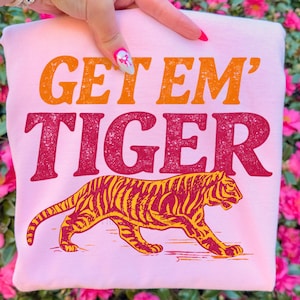 K&ouml;nnte beinhalten: Hellrosa Sweatshirt mit dem Text "GET 'EM TIGER" in Orange und Rot. Unter dem Text befindet sich eine detaillierte Illustration eines Tigers mit gelben und roten Streifen. Das Sweatshirt wird von einer Hand gehalten.