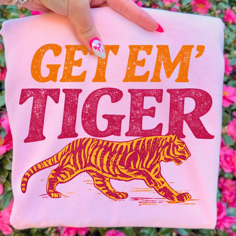 K&ouml;nnte beinhalten: Hellrosa Sweatshirt mit dem Text "GET 'EM TIGER" in Orange und Rot. Unter dem Text befindet sich eine detaillierte Illustration eines Tigers mit gelben und roten Streifen. Das Sweatshirt wird von einer Hand gehalten.
