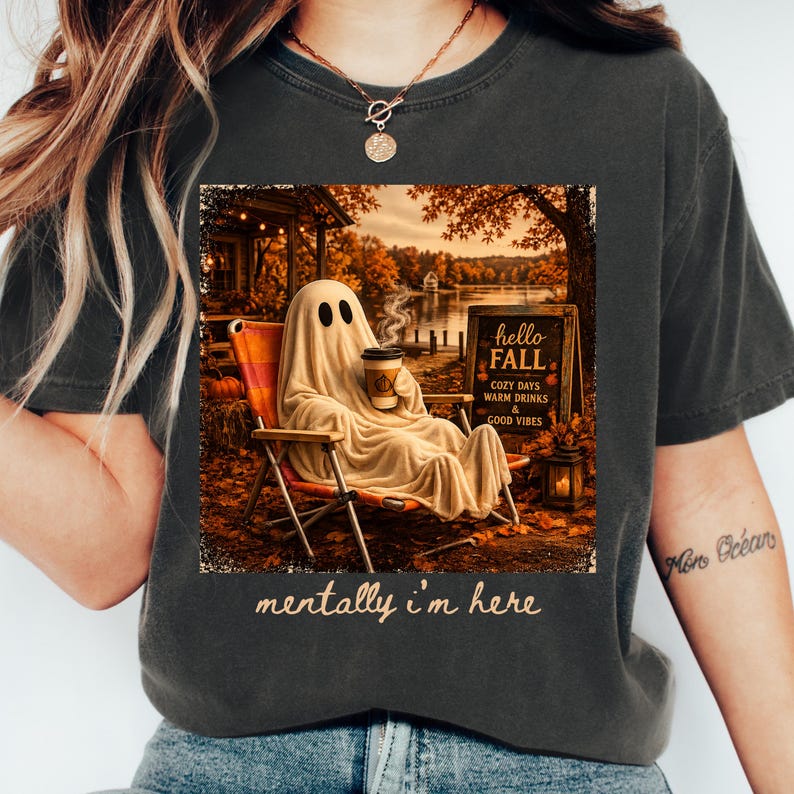 May include: Dark grey t-shirt featuring a fall-themed graphic. The graphic depicts a ghost in a chair holding a coffee cup, with the text "hello FALL" and "mentally i'm here". The background shows a lake and autumn foliage.