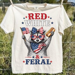 K&ouml;nnte beinhalten: Cremefarbenes T-Shirt mit einem Waschb&auml;ren, der eine herzf&ouml;rmige Sonnenbrille, eine Schleife und ein Halstuch in Rot, Wei&szlig; und Blau tr&auml;gt. Der Waschb&auml;r h&auml;lt eine amerikanische Flagge und einen Hotdog. Der Text auf dem Shirt lautet "RED WHITE & FERAL."