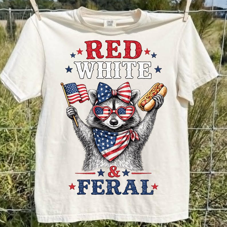K&ouml;nnte beinhalten: Cremefarbenes T-Shirt mit einem Waschb&auml;ren, der eine herzf&ouml;rmige Sonnenbrille, eine Schleife und ein Halstuch in Rot, Wei&szlig; und Blau tr&auml;gt. Der Waschb&auml;r h&auml;lt eine amerikanische Flagge und einen Hotdog. Der Text auf dem Shirt lautet "RED WHITE & FERAL."