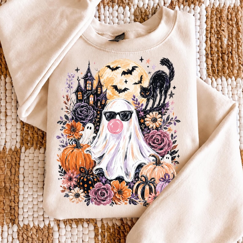 May include: A cream-coloured crewneck sweatshirt featuring a graphic print of a ghost wearing black sunglasses and blowing a pink bubble. The design includes a haunted house, a black cat, bats, a full moon, orange pumpkins, and purple flowers.