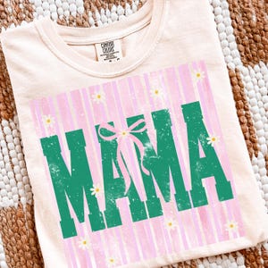 以下が含まれることがあります： 淡いピーチ色のTシャツ。色あせた緑色のブロック体で「MAMA」の文字がプリントされています。ピンクと白のストライプの背景、小さな白いデイジーのアクセント、ピンクのリボンがデザインされています。