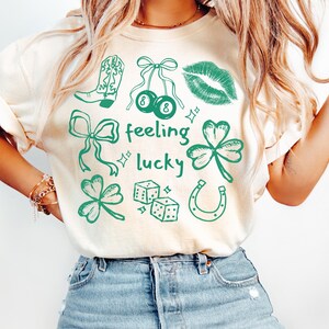 May include: Cream-colored t-shirt with a green graphic design featuring a cowboy boot, cherries, lips, bows, shamrocks, dice, and a horseshoe. The words "feeling lucky" are also printed on the shirt.
