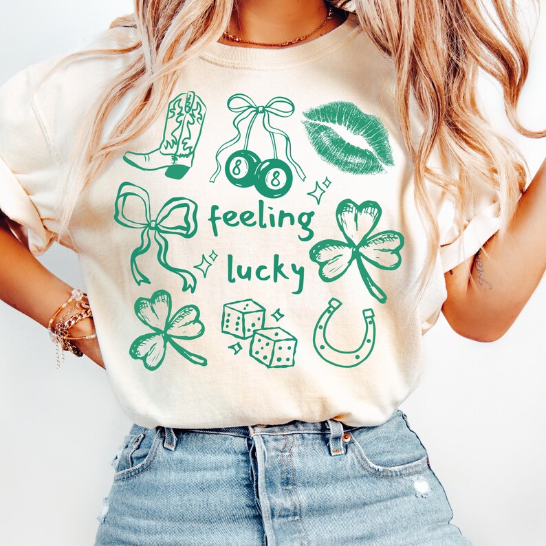 May include: Cream-colored t-shirt with a green graphic design featuring a cowboy boot, cherries, lips, bows, shamrocks, dice, and a horseshoe. The words "feeling lucky" are also printed on the shirt.