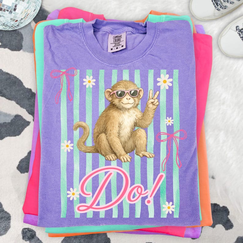 May include: A stack of colorful t-shirts. The top shirt is purple with a graphic of a monkey wearing sunglasses and giving a peace sign. The shirt has the word "Do!" in pink script. Other shirts are pink, orange, and green.