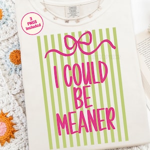 May include: Cream-colored t-shirt with the text "I COULD BE MEANER" in bright pink, set against a background of vertical green stripes. A pink bow design is above the text. The shirt includes a tag that says "Comfort Colors".