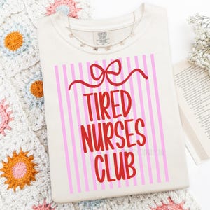 May include: Cream-colored t-shirt with the text "Tired Nurses Club" in red, set against a pink and white striped background. A red bow design is above the text. A delicate silver necklace with small charms is draped on the shirt.