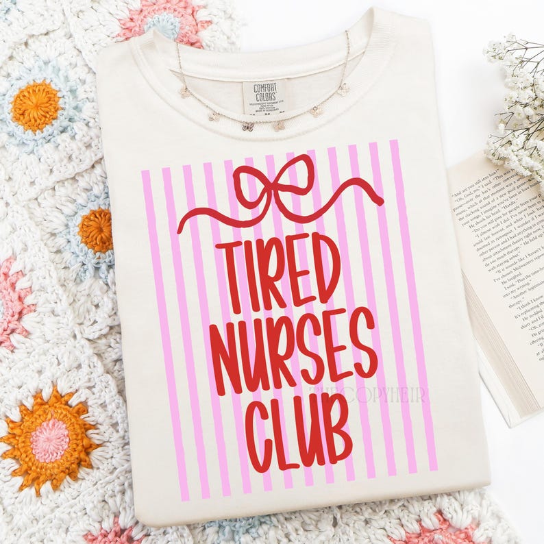 May include: Cream-colored t-shirt with the text "Tired Nurses Club" in red, set against a pink and white striped background. A red bow design is above the text. A delicate silver necklace with small charms is draped on the shirt.
