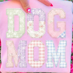 May include: A light pink sweatshirt with the words "DOG MOM" in large, patchwork-style letters. The letters are in various pastel colors and patterns, including floral and gingham designs.