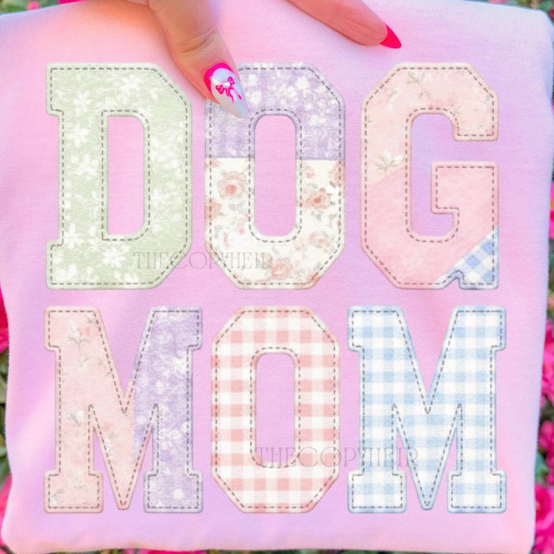 May include: A light pink sweatshirt with the words "DOG MOM" in large, patchwork-style letters. The letters are in various pastel colors and patterns, including floral and gingham designs.