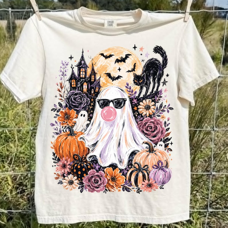 May include: A cream-coloured short-sleeve t-shirt featuring a graphic print of a ghost wearing sunglasses and blowing a pink bubble. The design includes a haunted house, a black cat, bats, a full moon, pumpkins, and various colourful flowers.