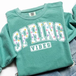 May include: A teal crewneck sweatshirt with the words "SPRING VIBES" in a white, floral-filled font. The sweatshirt is folded on top of a pair of blue jeans.