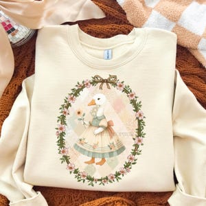 May include: Cream-colored sweatshirt featuring a whimsical illustration of a goose wearing a dress and holding a flower, framed by a floral wreath. The design includes a bow and the text "THEOPHEIR".