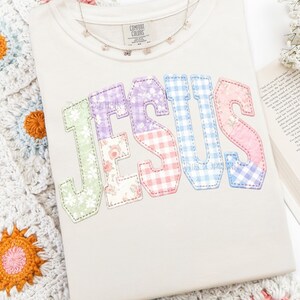 Puede incluir: Sudadera color crema con la palabra "JESUS" aplicada en telas de patchwork coloridas. Las letras presentan estampados florales, cuadros y lisos en tonos pastel. Un delicado collar de mariposas descansa sobre la camisa.