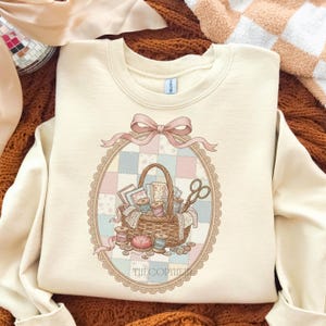 May include: Cream-colored sweatshirt featuring a detailed illustration of a sewing basket filled with spools of thread, fabric scraps, and scissors. The design is framed by a pink bow and an oval border with a patchwork background. The text "THECOPYHER" is at the bottom.