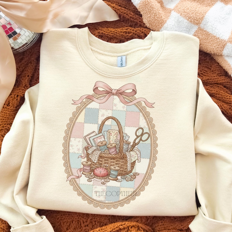 May include: Cream-colored sweatshirt featuring a detailed illustration of a sewing basket filled with spools of thread, fabric scraps, and scissors. The design is framed by a pink bow and an oval border with a patchwork background. The text "THECOPYHER" is at the bottom.