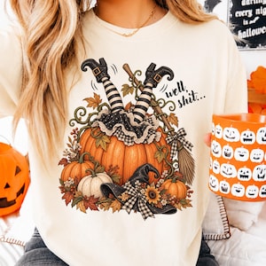 May include: A cream-colored t-shirt featuring a graphic of a large orange pumpkin with witch legs in black and white striped stockings sticking out of the top. The design includes a broom, a black witch hat, autumn leaves, and the text well, shit...