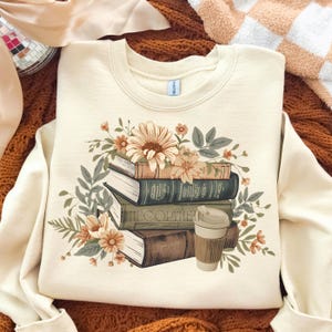 May include: Cream-colored sweatshirt featuring a stack of books with floral accents and a coffee cup. The books are in shades of brown and green, with a large flower at the top. The text "THE COPYHER" is visible on one book.