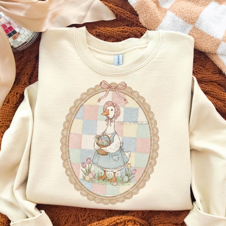 Puede incluir: Sudadera color crema con una ilustraci&oacute;n de un ganso con gorro y delantal, sosteniendo una cesta de huevos de Pascua. El ganso est&aacute; enmarcado en un &oacute;valo con un fondo de retazos. El texto "THE COPIER" es visible.
