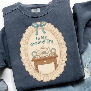 May include: A navy blue sweatshirt with a graphic featuring a tea set on a table, inside an oval frame with the text "In My Granny Era". The frame is adorned with a blue bow and a ruffled border.