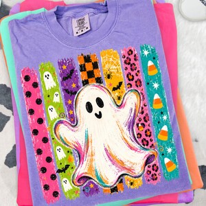 May include: A purple short-sleeve t-shirt featuring a large white ghost graphic centred over vertical brushstroke patterns. The patterns include black polka dots, white ghosts, bats, orange and black checkers, spiderwebs, leopard print, and candy corn.