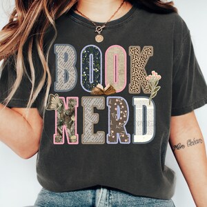 May include: Dark gray t-shirt with the words "BOOK NERD" in a colorful, patchwork design. The letters are filled with patterns like floral, leopard print, and book pages. A butterfly and a small open book are also featured.