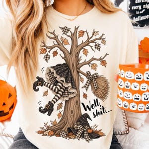 May include: A cream-colored t-shirt featuring a graphic of a tree with a witch's legs and broomstick stuck in the branches. The design includes a black witch hat, falling autumn leaves, and the text "Well shit..." in a black script font.