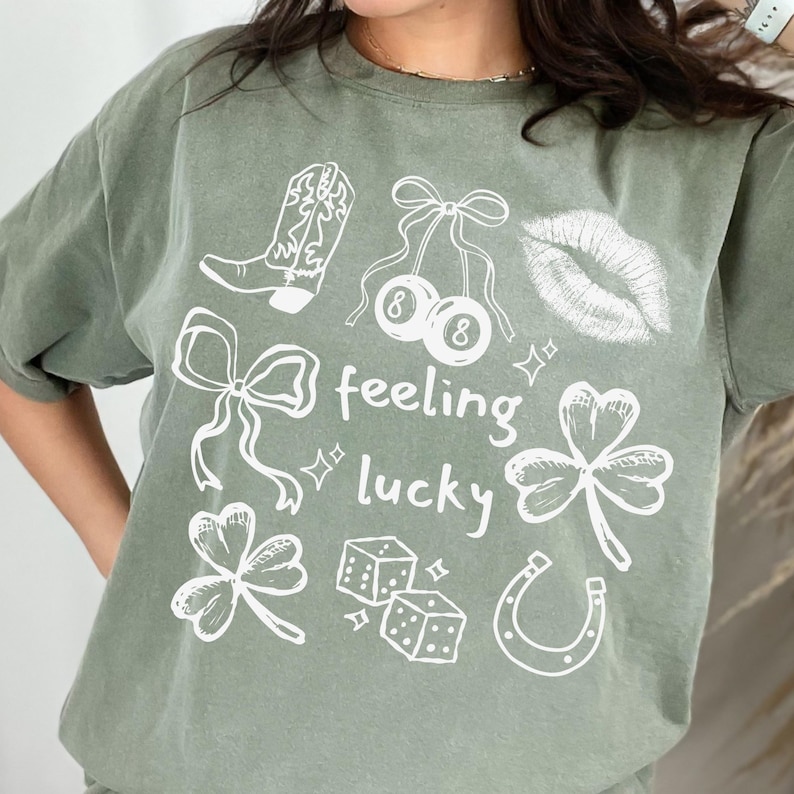 May include: Sage green t-shirt with white graphic illustrations of a cowboy boot, cherries, lips, bows, shamrocks, dice, and a horseshoe. The words "feeling lucky" are written in a playful font.