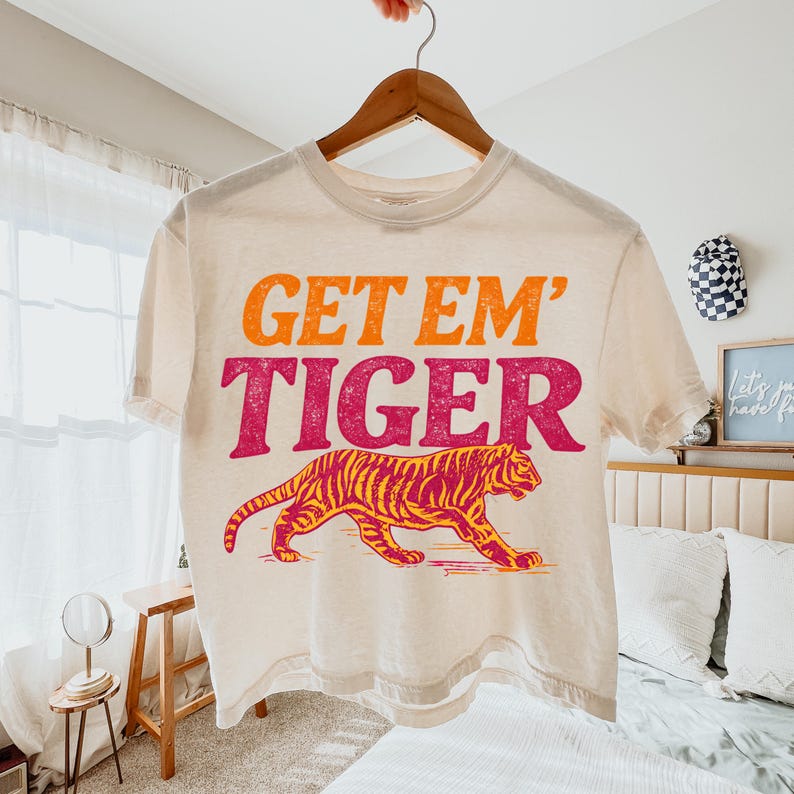 May include: Cream-coloured t-shirt with the text "GET EM' TIGER" in orange and pink. A tiger illustration in orange and yellow is below the text. The shirt is hanging on a wooden hanger.