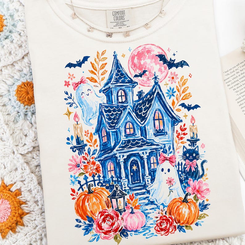 May include: A cream-colored t-shirt featuring a colorful graphic print of a blue haunted house, two ghosts with pink bows, a black cat, orange pumpkins, flowers, and bats under a pink moon. The shirt has a Comfort Colors label at the neckline.