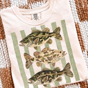 May include: A light pink t-shirt featuring three detailed fish illustrations in shades of green, brown, and tan. Vertical green stripes are printed on the shirt. The brand "Comfort Colors" is printed on a tag at the neckline.