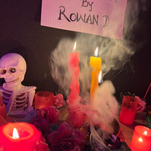 May include: A mystical scene featuring lit candles, a skeleton figurine, and floral arrangements. The image includes a sign that reads "Moonchant by Rowan D." Red and yellow candles are burning, with smoke rising. The scene is set on a wooden surface.