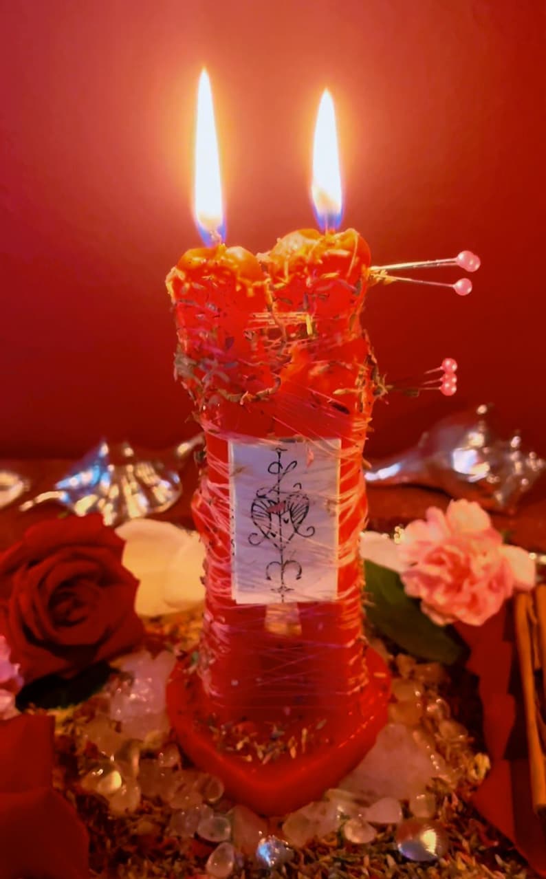 May include: A red candle with two flames, decorated with twine, herbs, and a small label with a heart symbol. The candle is surrounded by flowers, crystals, and a red background, creating a mystical ambiance.