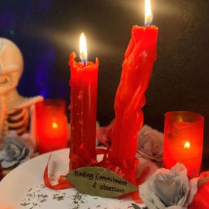May include: Two red wax candles with lit flames, one shaped like a human figure, tied with red ribbon. A dried bay leaf rests in front with the handwritten text Binding Commitment & obsession. The candles are set on a white plate with grey flowers.
