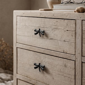 May include: A light-coloured wooden chest of drawers with three drawers. The chest has black dragonfly-shaped drawer pulls. A vase with dried flowers, books, and spectacles are on top.
